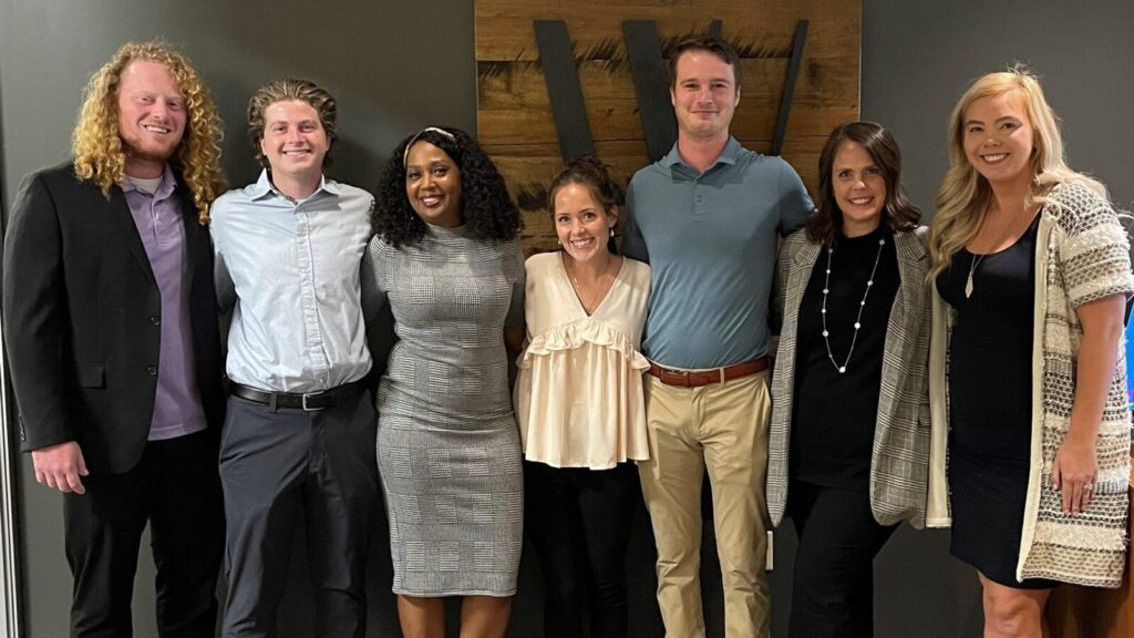 Seven people standing side by side indoors, posing for a group photo in business casual and professional attire, with a wood panel and logo in the background.