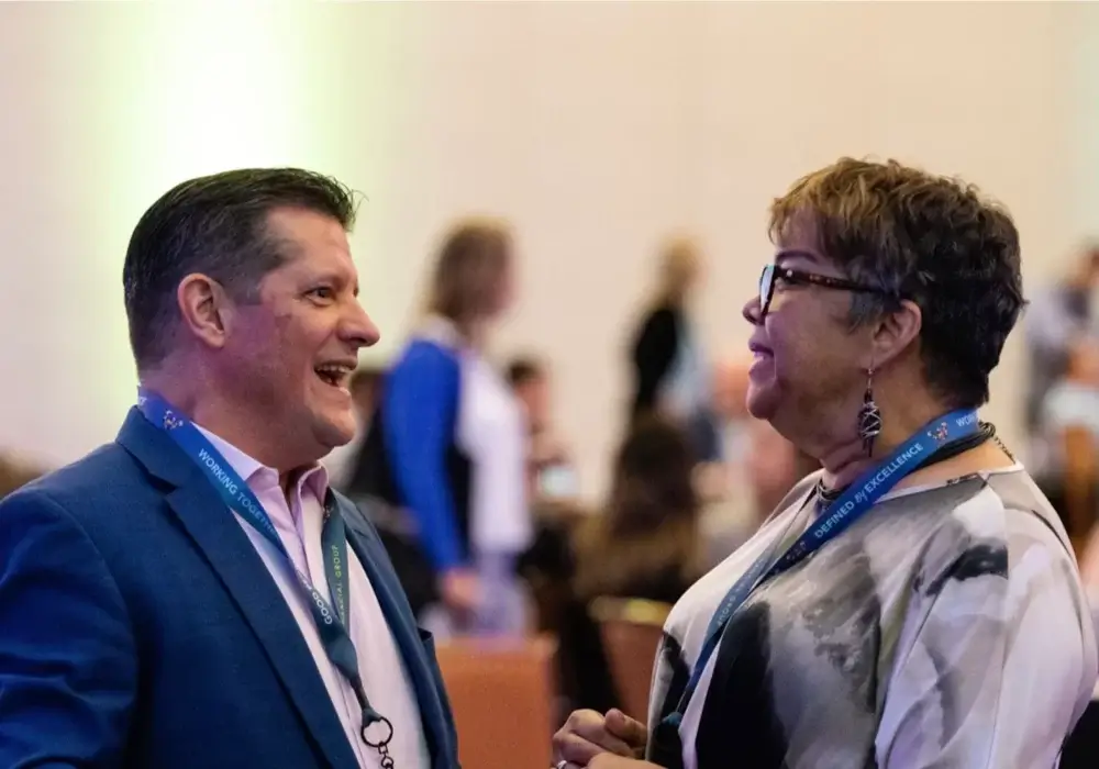 A man and a woman wearing conference badges are standing and talking to each other in a crowded indoor setting.