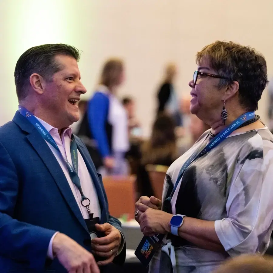 Two people wearing conference badges and lanyards are standing and having a conversation at an indoor event, with other attendees visible in the background.