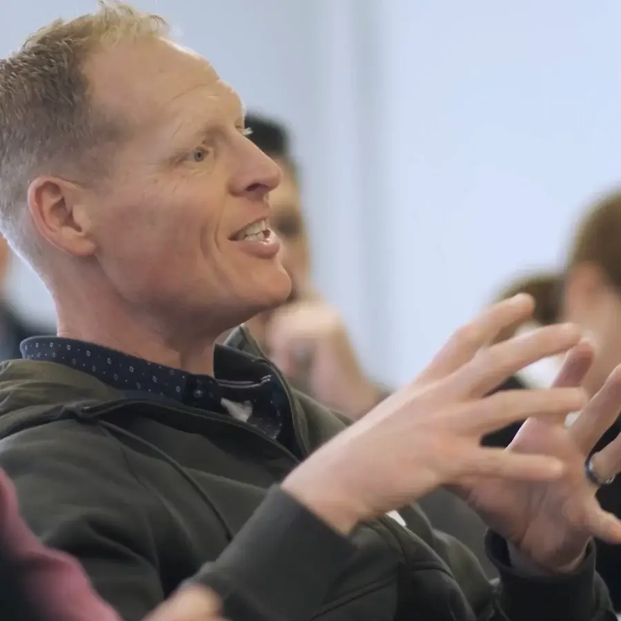 A man with short light hair gestures with both hands while speaking in what appears to be a group setting or discussion.