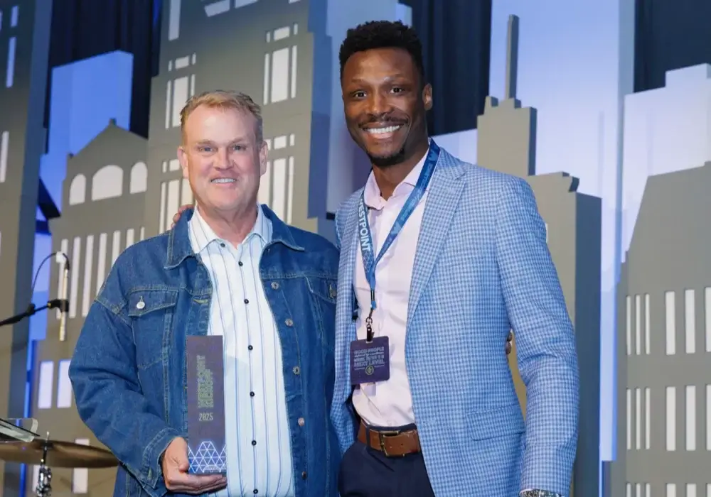 Two men stand side by side at an event; one holds an award, and both are smiling in front of a backdrop with building silhouettes.