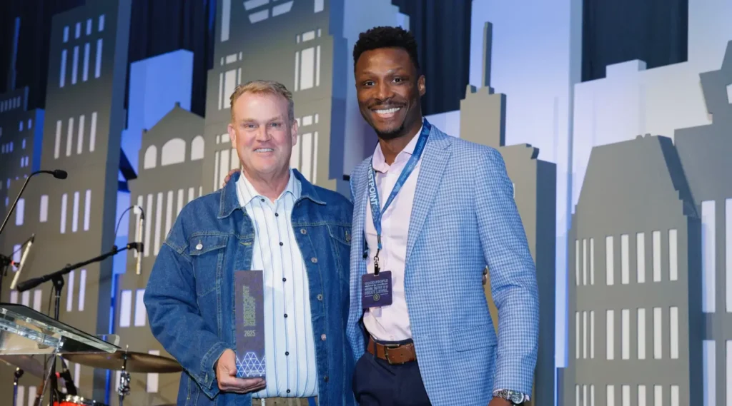 Two men stand side by side at an indoor event; one holds a trophy. Both are smiling. A cityscape-themed backdrop is behind them.