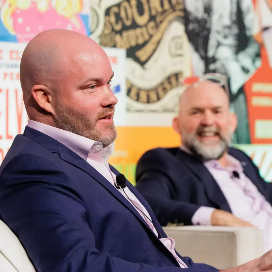 Two men in suits with microphones sit and talk on a stage in front of a colorful, patterned backdrop.
