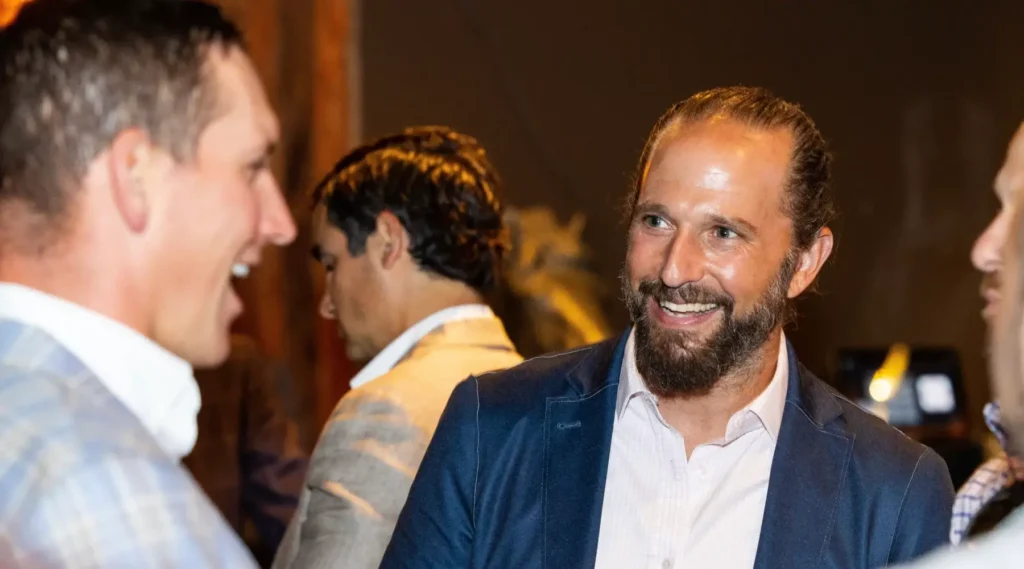 A group of men in business attire engage in conversation at an indoor event. One man with a beard and ponytail smiles while talking to another man.