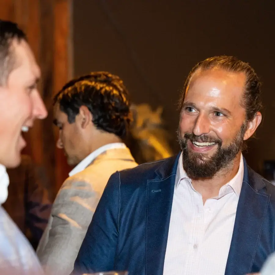 Two men in suits are talking and smiling at an indoor event, with another person in the background facing away.
