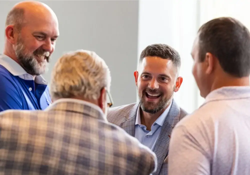 Four men in business attire stand together indoors, smiling and talking in a casual, brightly lit setting.