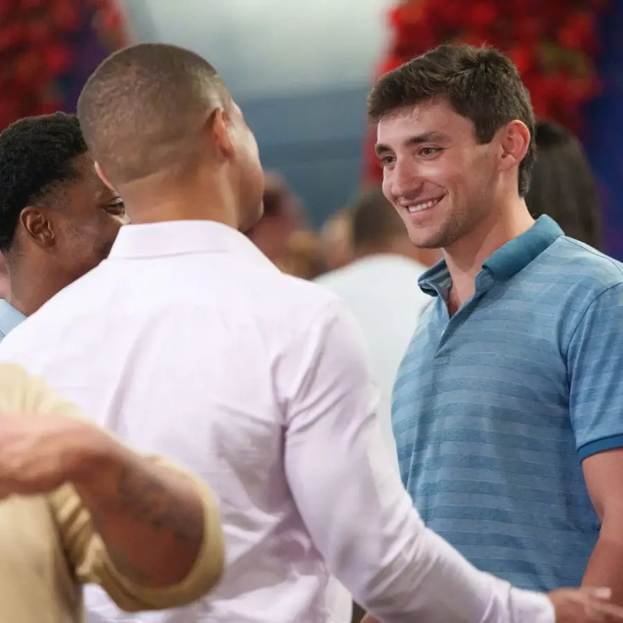 Two men stand facing each other and smiling while having a conversation at an indoor social event with other people in the background.