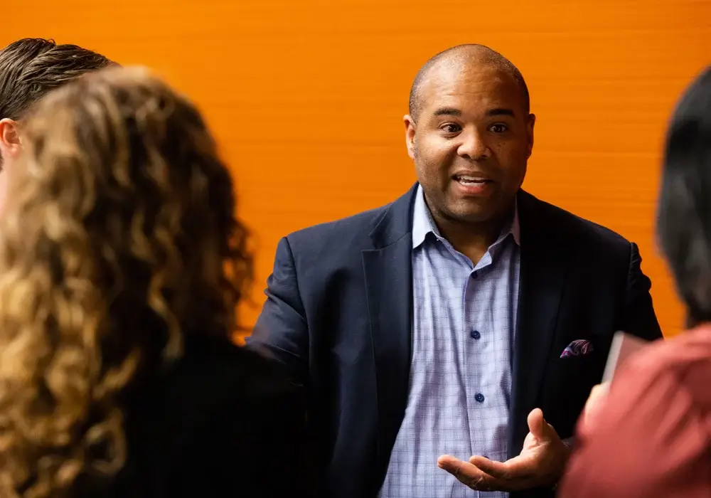 A man wearing a dark blazer and checked shirt speaks to a small group of people in front of an orange background.