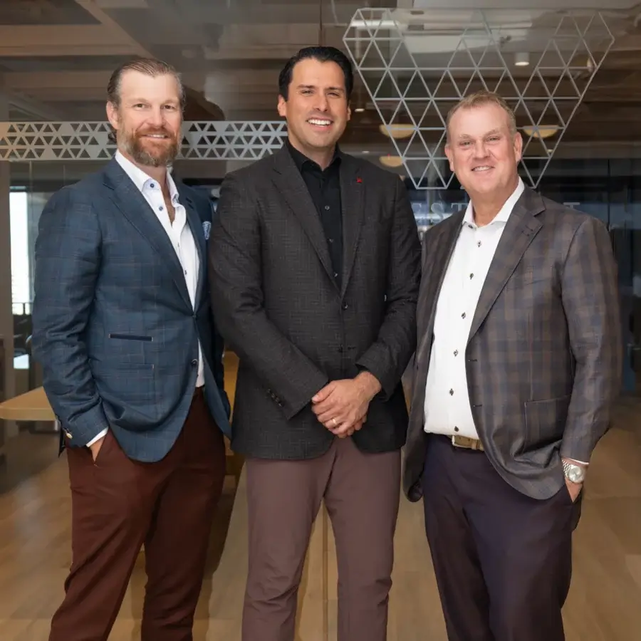 Three men wearing business attire stand together in an office setting, smiling at the camera.