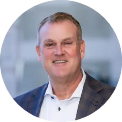 A middle-aged man with short light hair, wearing a white shirt and a dark checkered blazer, smiling in front of a blurred indoor background.