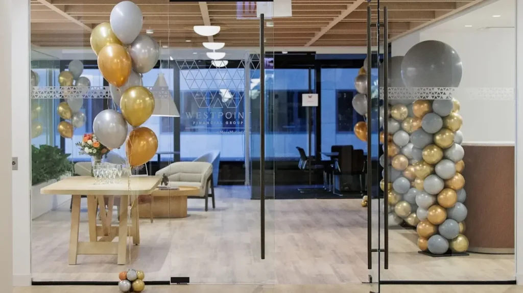 Office entrance decorated with gold, silver, and gray balloons, a table with glasses and drinks, and seating visible through glass doors.