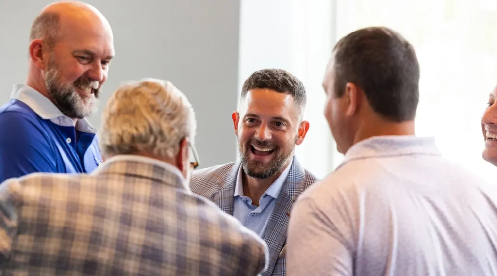 Five men in collared shirts and blazers stand in a group indoors, engaged in conversation and smiling.