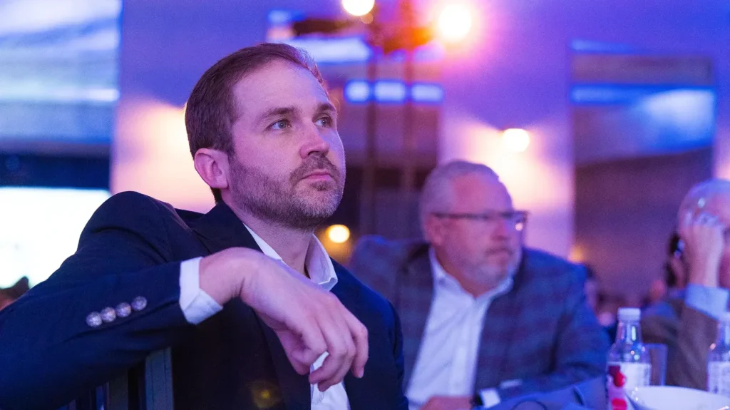 A man in a suit sits at a table, attentively watching something off-camera, with other people and blue lighting in the background.