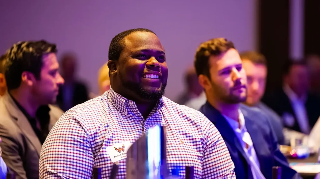 A man in a checkered shirt smiles while seated among other attendees at an indoor event or conference.