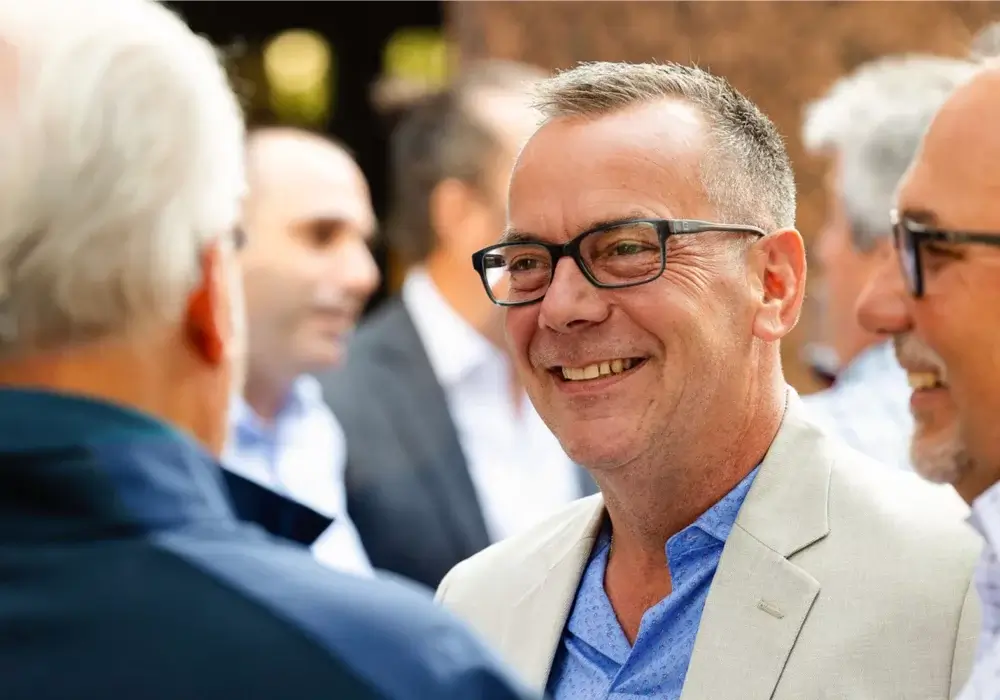 A group of men in business attire stand together outdoors, with one man in the center smiling and wearing glasses.
