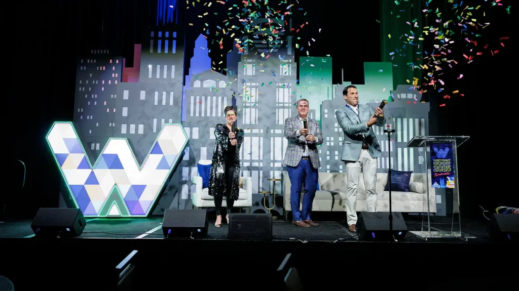 Three people on stage celebrate with confetti poppers in front of a cityscape backdrop and a large geometric "W" sign.