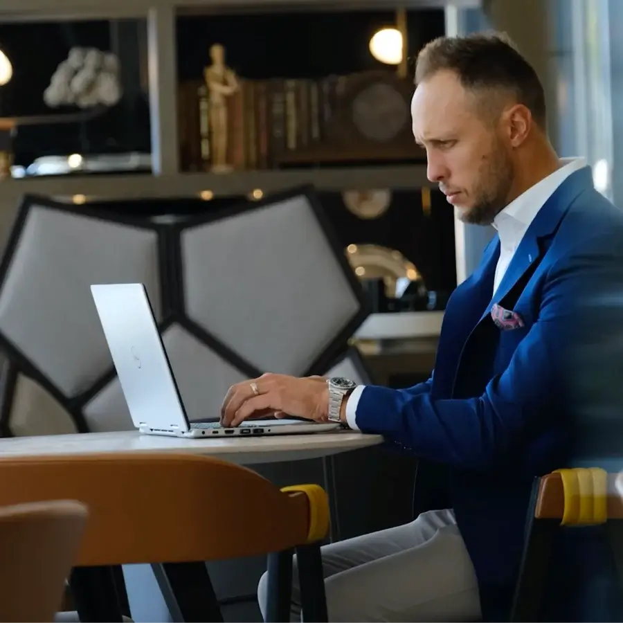 A man in a blue suit works on a laptop at a table in a modern, well-lit indoor setting with shelves and decor in the background.