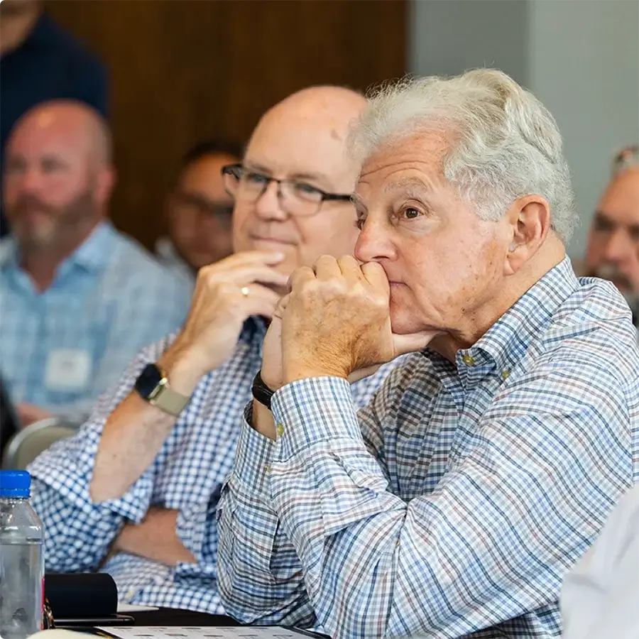 Two older men sit attentively at a meeting or conference, both wearing glasses and plaid shirts, with other people blurred in the background.