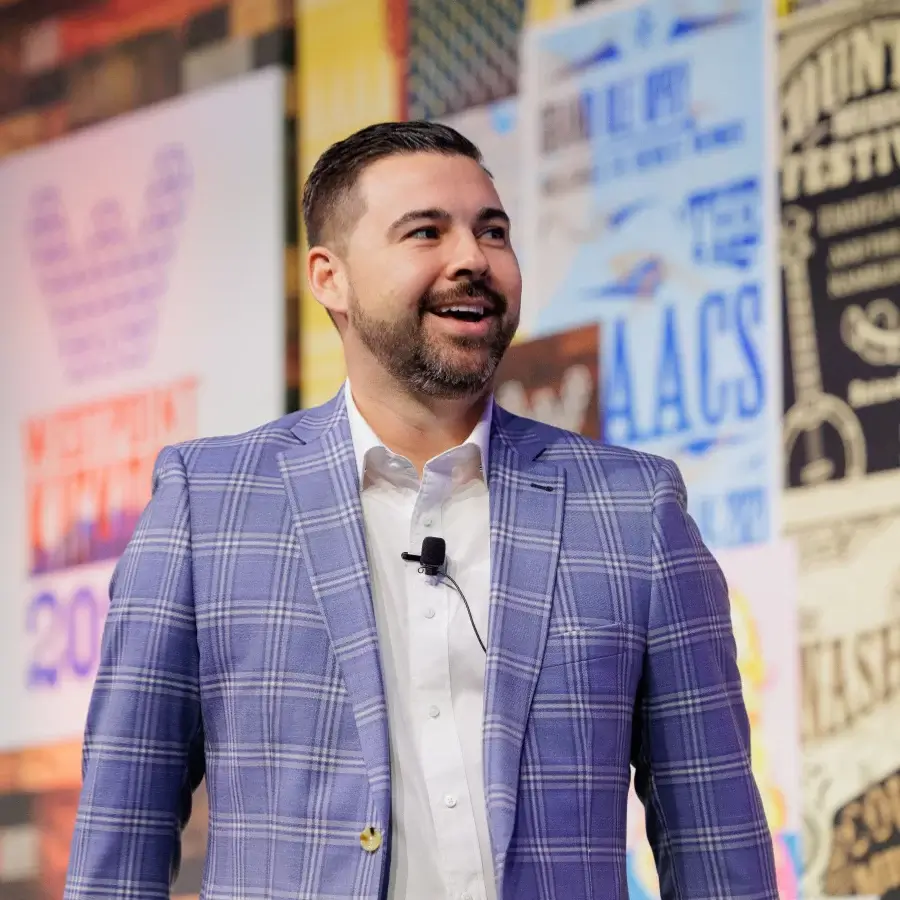 A man in a checked blazer and white shirt stands smiling, wearing a microphone headset, with colorful posters displayed on the wall behind him.