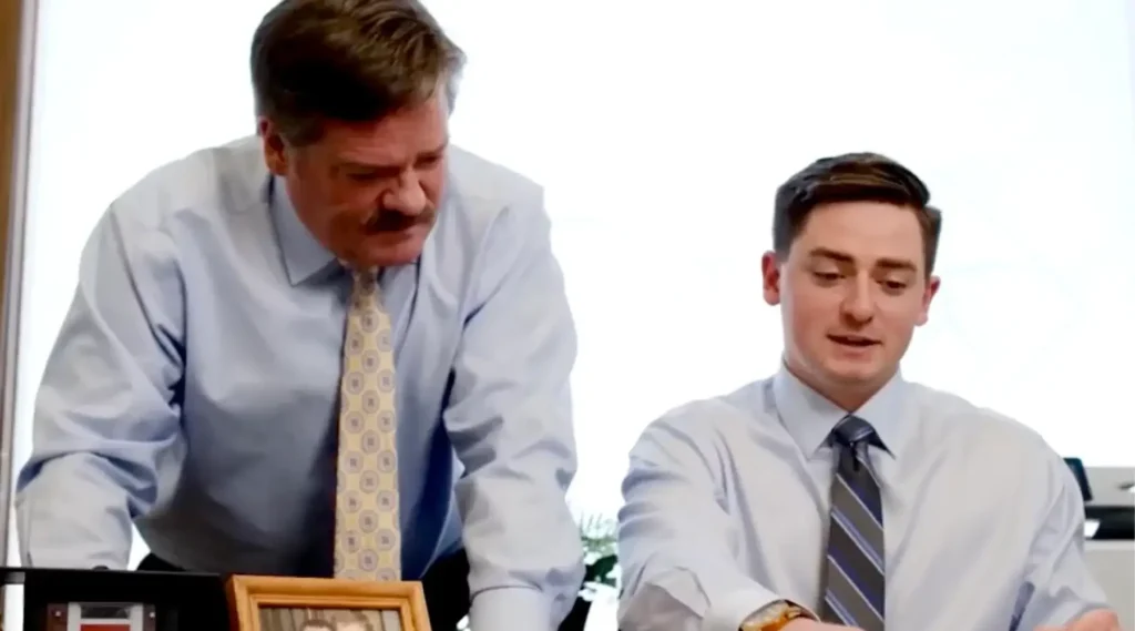 Two men in dress shirts and ties are working together at a desk, with one standing and leaning over while the other is seated. A framed photograph is visible on the desk.
