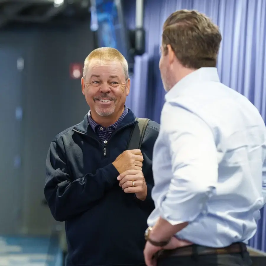 Two men stand indoors having a conversation; one is smiling and holding a strap over his shoulder, while the other stands with his back to the camera.