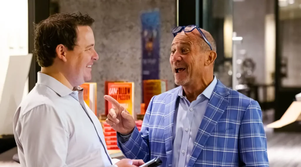 Two men stand indoors, smiling and talking. One man holds a phone while the other gestures with his finger, wearing glasses on his head and a blue plaid jacket. Books are visible in the background.