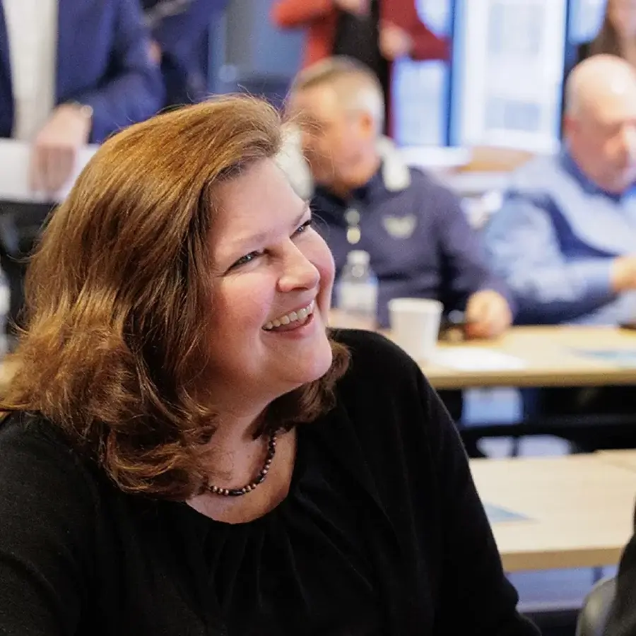 A woman with brown hair smiles while seated at a table in a conference room with other people in the background.
