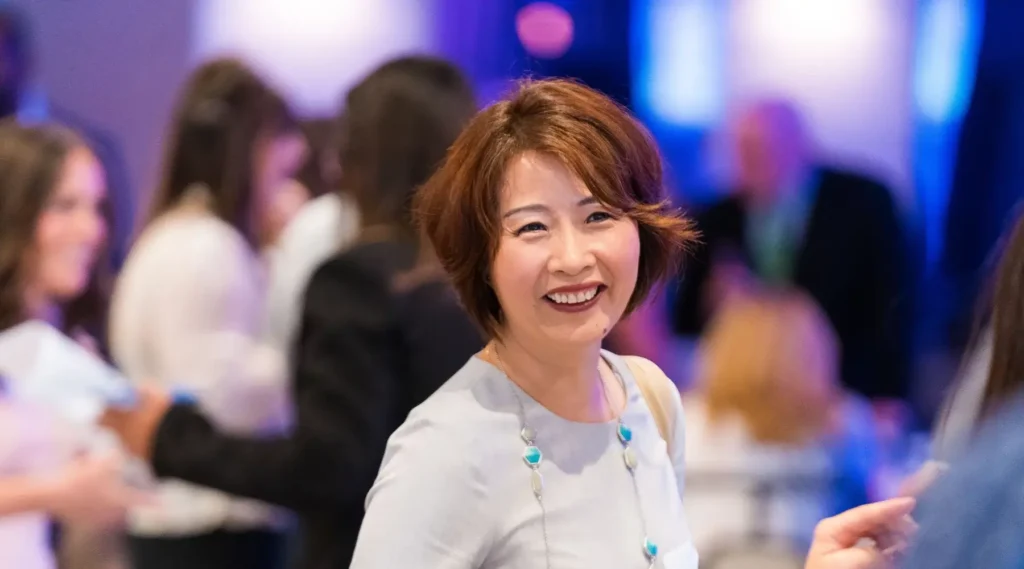 A woman with short brown hair and a light-colored dress smiles while standing in a crowded indoor event space with people in the background.