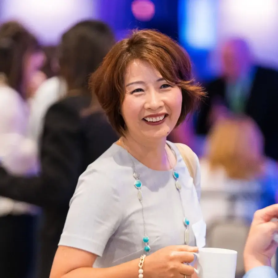 A woman with short brown hair smiles while holding a white cup at a social event, with people blurred in the background.