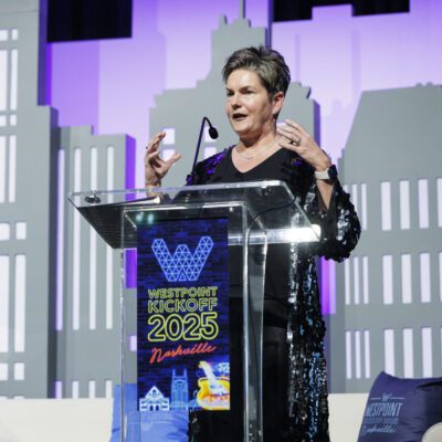 A woman speaks at a podium labeled "WestPoint Kickoff 2025 Nashville" on a stage with cityscape-themed decorations in the background.
