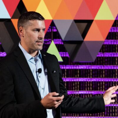 A man in a suit jacket speaks on stage, gesturing with one hand, with a microphone clipped to his shirt and a colorful geometric backdrop behind him.