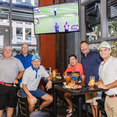 Six men pose inside a restaurant, some seated and some standing around a table with drinks and snacks. A golf tournament is shown on a TV mounted above them.