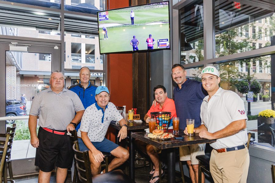 Six men pose inside a restaurant, some seated and some standing around a table with drinks and snacks. A golf tournament is shown on a TV mounted above them.