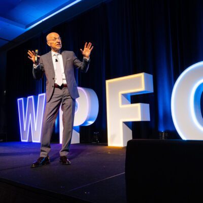 A man in a suit speaks on stage with large illuminated letters "WPFG" behind him and a podium nearby.