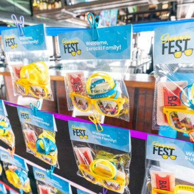 Rows of clear plastic bags containing yellow sunglasses, wristbands, and cards hang on clips from a display at an indoor event labeled "Summer FEST.
