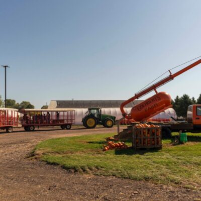 A tractor pulls passenger wagons near a large orange cannon labeled "Ciderbits Family," with pumpkins displayed on the grass and a truck parked nearby.