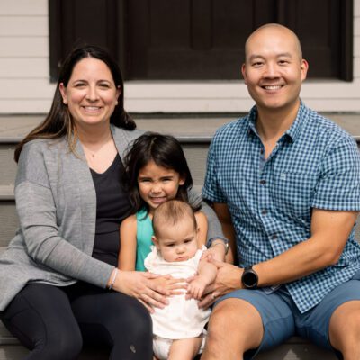 A man, woman, young girl, and baby sit together on outdoor steps, posing for a family photo in daylight.