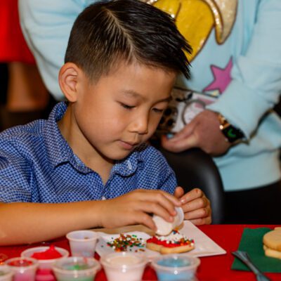A young boy decorates cookies with frosting and sprinkles at a table, with various containers of colorful toppings around him.