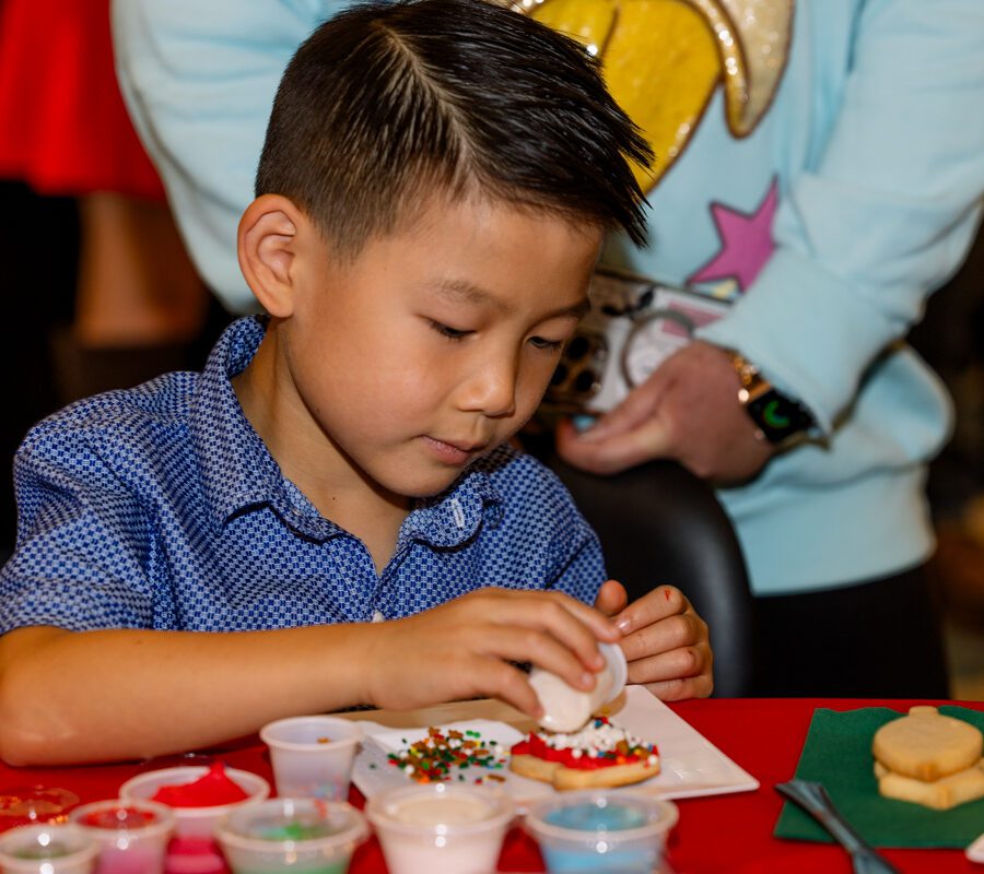 A young boy decorates cookies with frosting and sprinkles at a table, with various containers of colorful toppings around him.