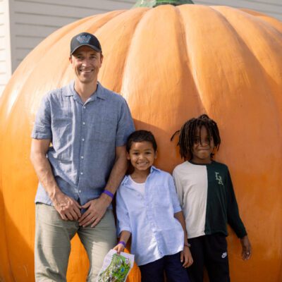 A man and two children stand and smile in front of a large orange pumpkin outdoors.