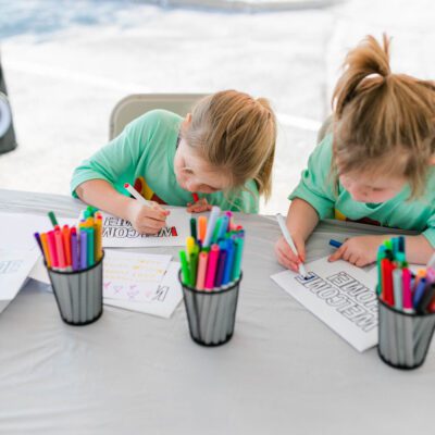 Two young children sit at a table coloring printed sheets with markers, surrounded by containers filled with colorful markers.