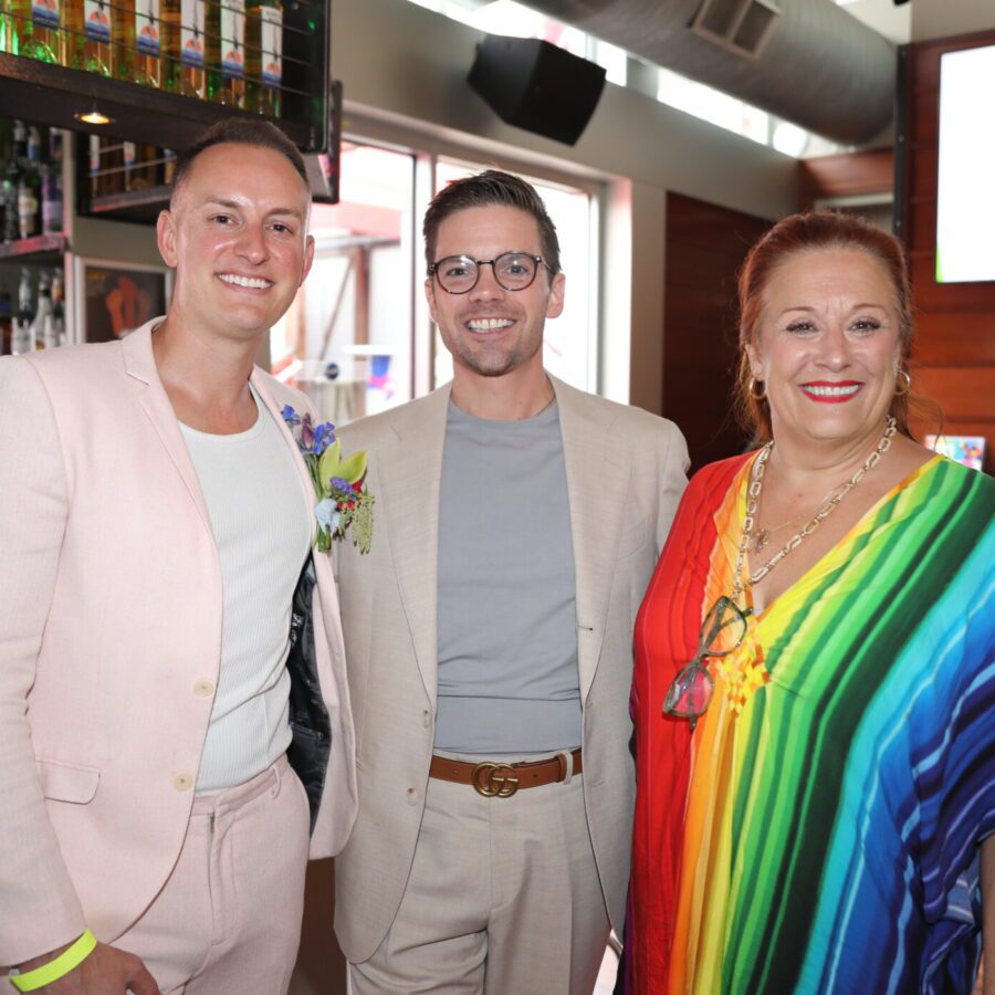 Three people pose together in a bar; two men wear light-colored suits and a woman wears a rainbow-striped dress. A screen in the background displays "PRIDE PARTY.