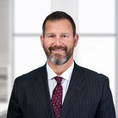 A man with short brown hair and a beard, wearing a dark pinstripe suit, white shirt, and maroon polka dot tie, stands in front of a blurred office background.