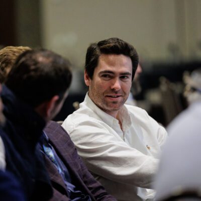 A man in a white shirt sits among a group of people in conversation at an indoor event.