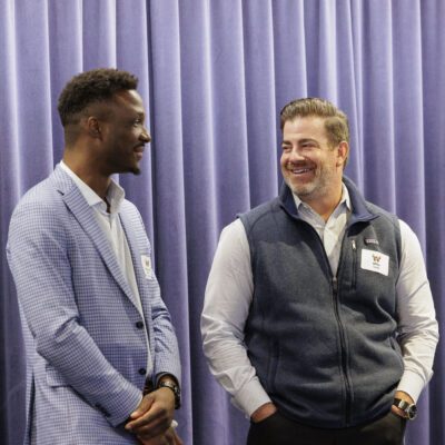 Two men stand in front of purple curtains, smiling and talking. One wears a blue checked suit, and the other wears a gray vest over a white shirt. Both have name tags.