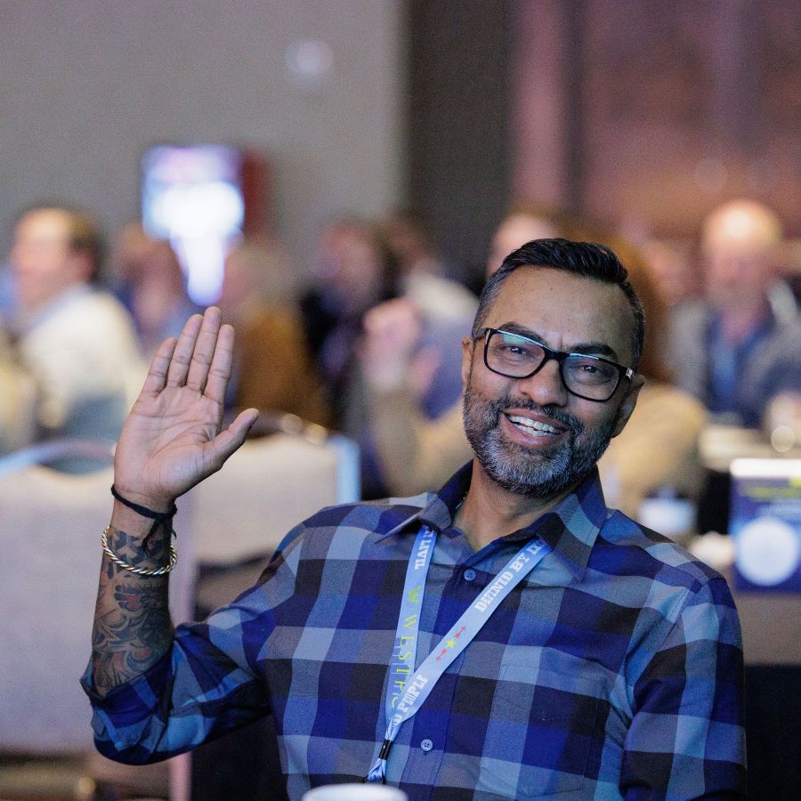 A man wearing glasses and a blue plaid shirt smiles and waves while seated at an indoor event with other attendees in the background.