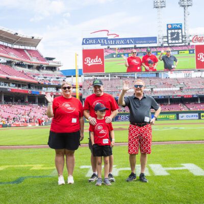 Four people in Reds attire stand on a baseball field at Great American Ball Park, with a large scoreboard and stadium seating visible in the background.
