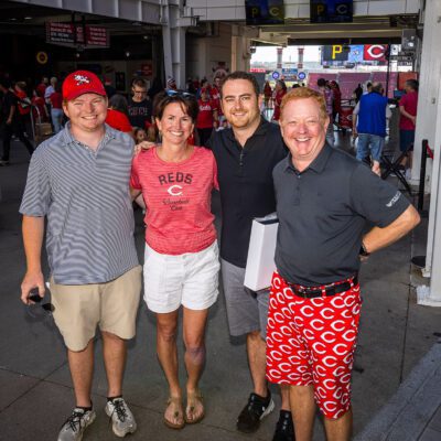 Four people pose together at a baseball stadium entrance, with two wearing Reds apparel and one in red "C" patterned shorts; other fans and concessions are visible in the background.