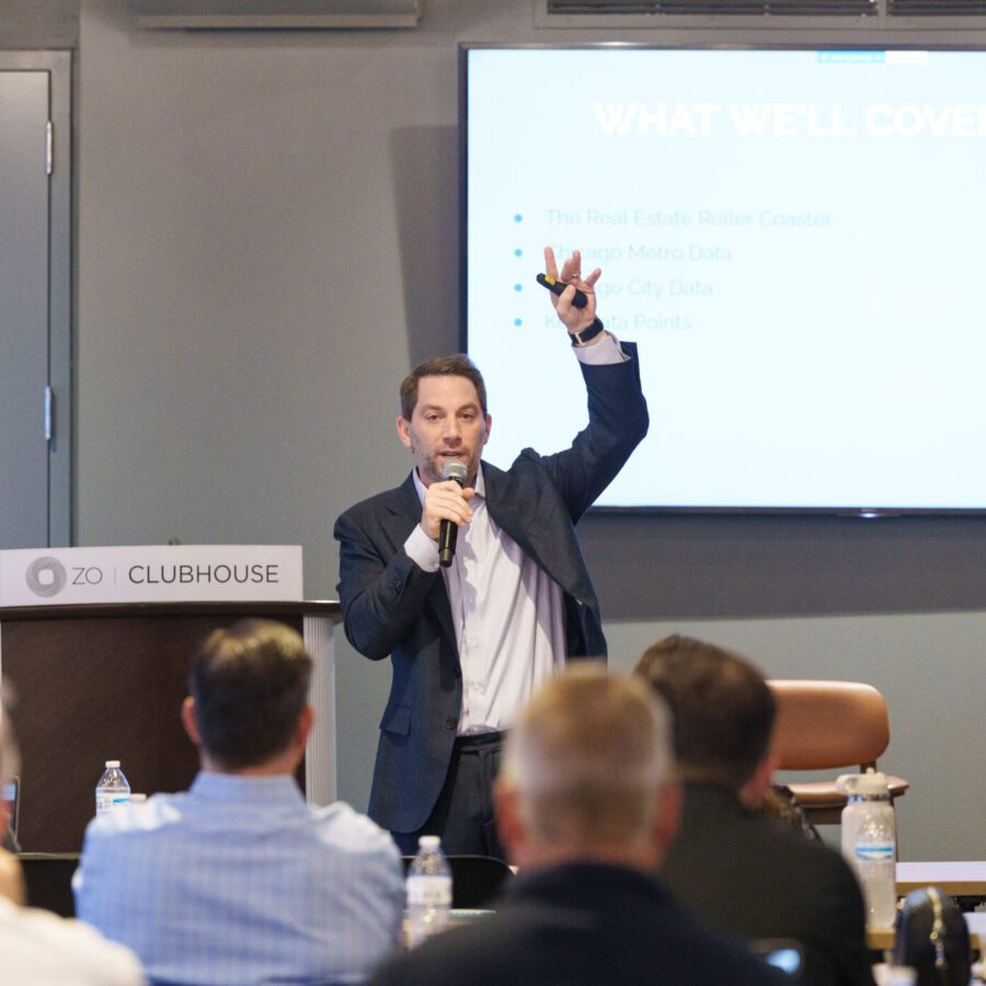 A man in a suit speaks into a microphone and gestures with his hand during a presentation to an audience in a conference room. A slide titled "WHAT WE'LL COVER TODAY" is displayed behind him.