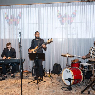 Three musicians perform indoors; one plays keyboard, one plays bass guitar, and one plays drums. A logo reading "West Point" is displayed on the curtain behind them.
